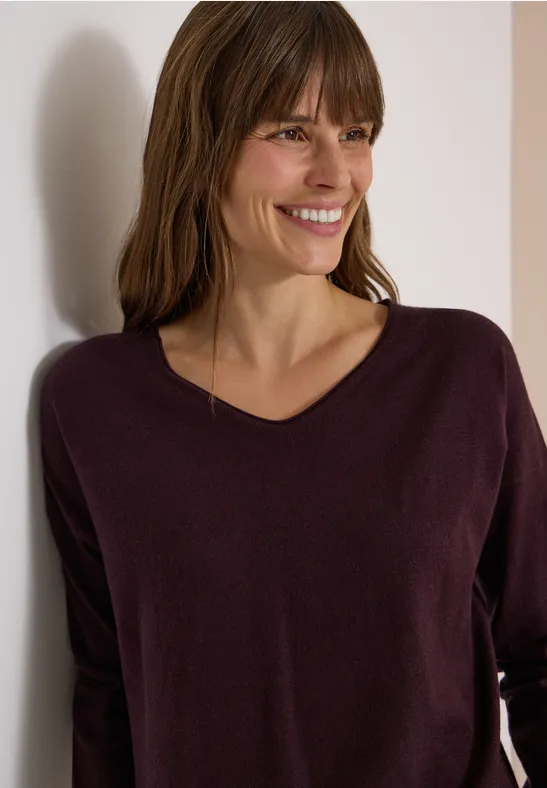 Eine lächelnde Frau in einem pflaumenfarbenen V-Ausschnitt-Pullover, angelehnt an eine weiße Wand.
