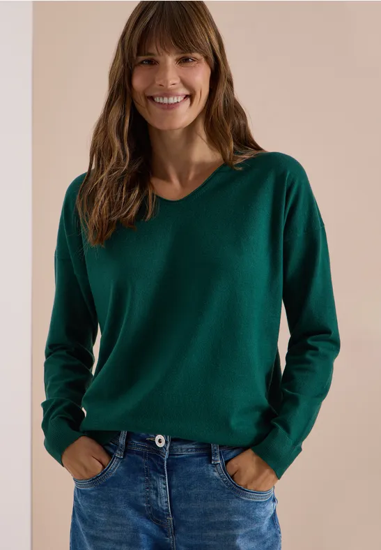 Eine lächelnde Frau in grünem Pullover und Jeans vor neutralem Hintergrund.