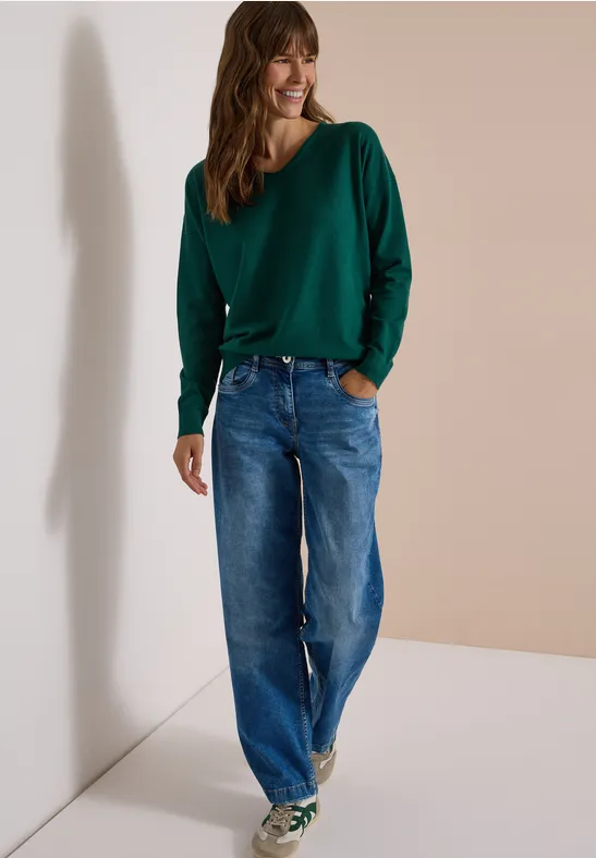 Eine junge Frau trägt einen grünen Pullover, weite blaue Jeans und grüne Sneaker vor neutralem Hintergrund.