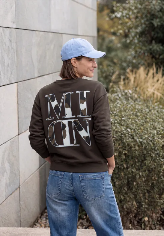 Frau an Mauer lehnt, trägt Jeans, Baseballkappe und dunklen Pullover mit markantem Rückendruck.