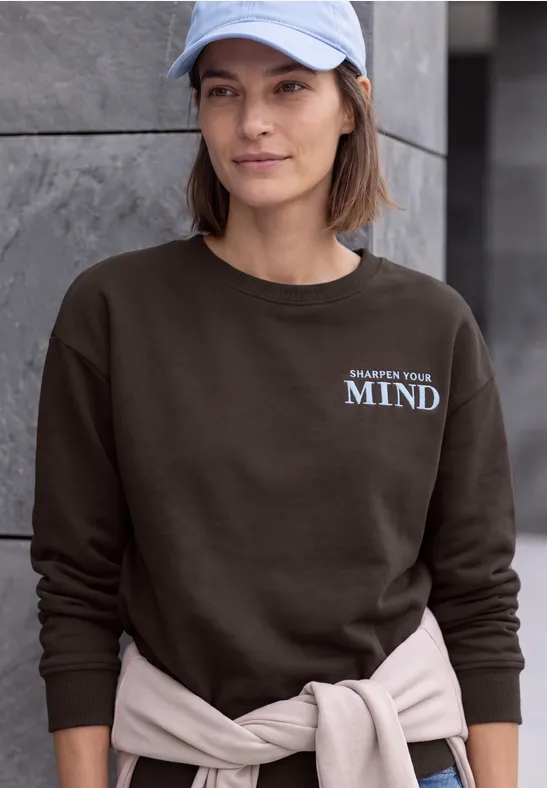 Frau mit hellblauer Cap, dunkelbraunem Sweatshirt mit Aufdruck 'A calmer mind' und hellem Pullover um die Taille