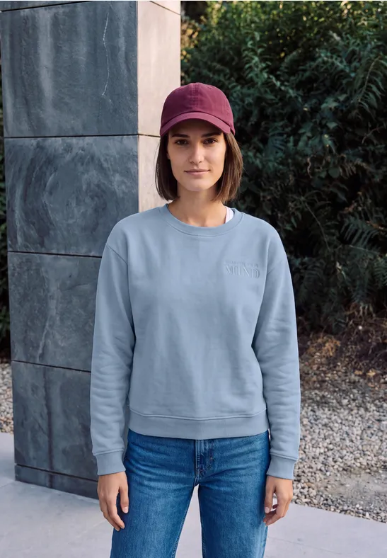 Frau steht vor Steinwand im Garten, trägt hellblaues Sweatshirt, Jeans und rotes Cap.