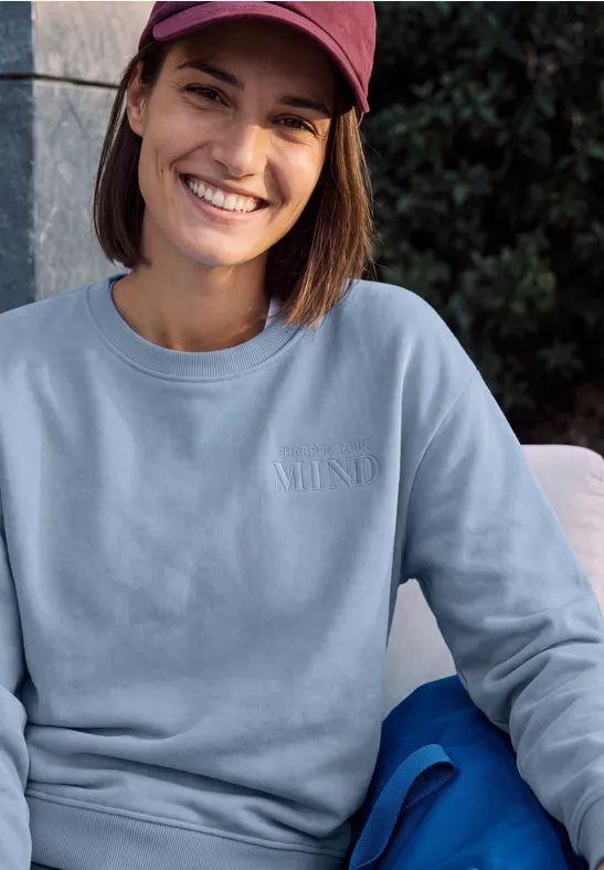 Junge Frau mit roter Baseballkappe und hellem Sweatshirt sitzt lächelnd vor Grünpflanzen.