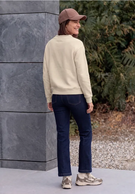 Frau in Sweatshirt, Jeans, Sneakern und Cap steht im Freien mit dem Rücken zur Kamera, schaut seitlich.