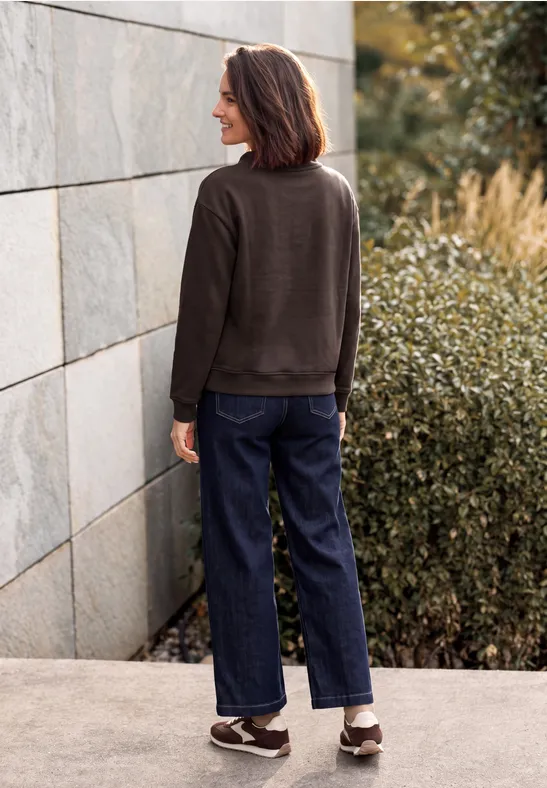 Frau von hinten in lässigem Outfit (Pullover, Jeans, Sneakers) steht vor einer Steinwand und Büschen.