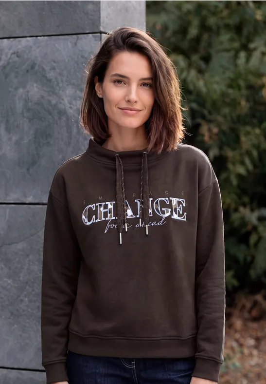 Eine lächelnde Frau im braunen Hoodie steht vor einer Steinwand und grünen Sträuchern.