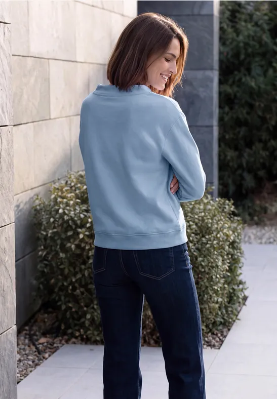 Frau in blauem Pullover und Jeans lehnt seitlich an Hauswand, Heckengebüsch im Hintergrund.
