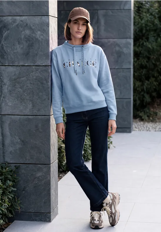 Frau in blauem Sweatshirt, Jeans, Sneakern und Cap lehnt an moderner Steinwand im Freien.