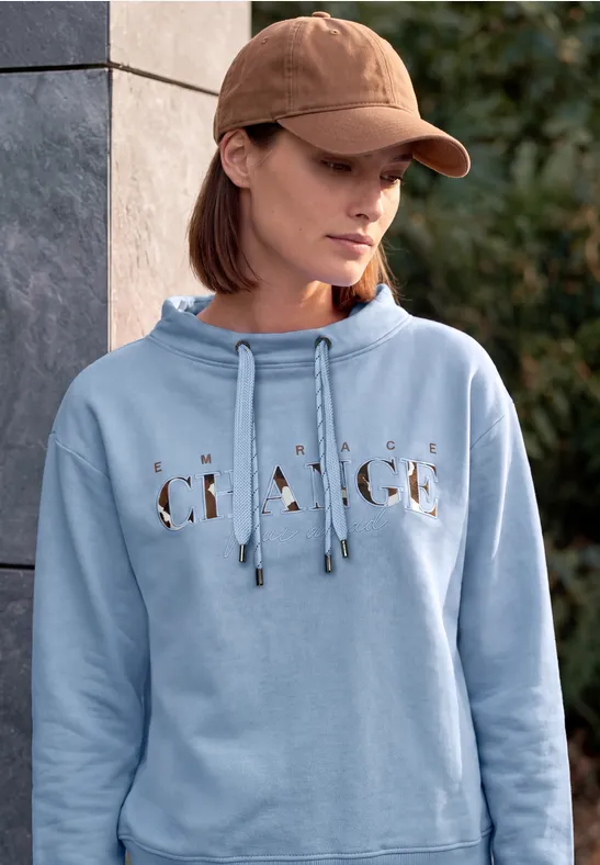 Frau in hellblauem Hoodie mit Schriftzug und Baseballcap lehnt entspannt an einer Steinsäule im Freien.