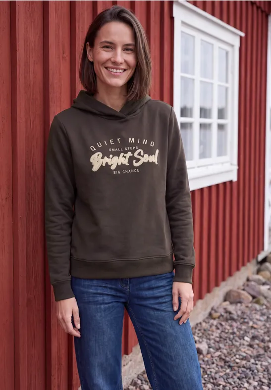 Een lachende vrouw voor een rood houten huis, draagt jeans en een hoodie met 'Bright Soul'.