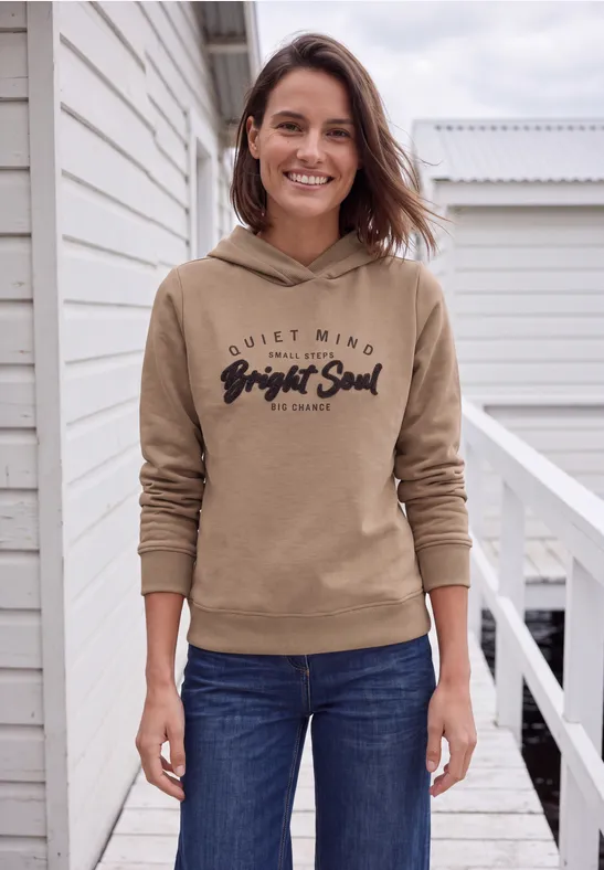 Frau mit brauner Kapuzenjacke und Jeans auf einer Veranda, lächelnd.