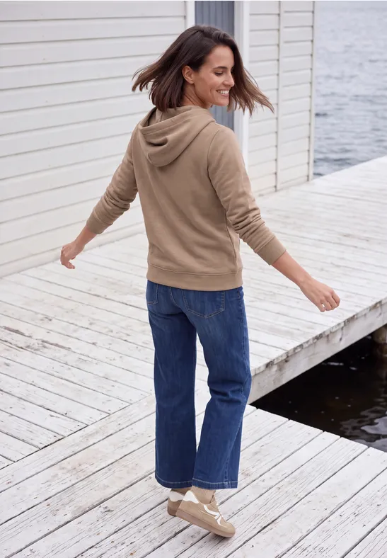 Eine Frau in Kapuzenpullover und Jeans dreht sich fröhlich auf einem Holzsteg am Wasser.