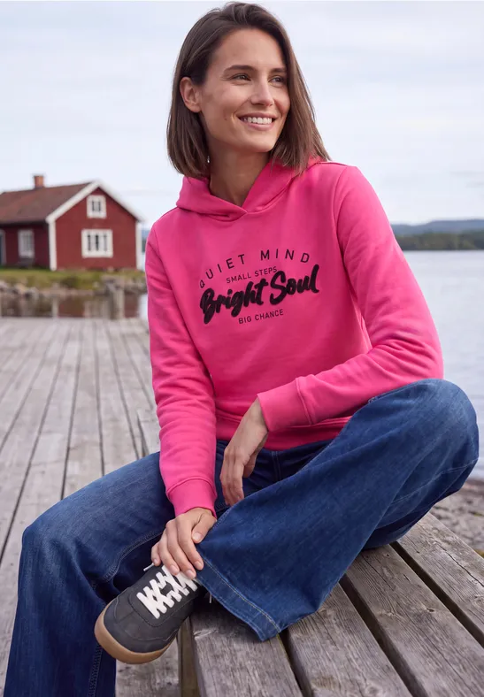 Frau in pinkem Hoodie sitzt auf Holzsteg am See und lächelt entspannt.