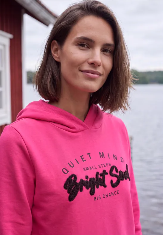 Junge Frau in pinkem Hoodie steht lächelnd am Seeufer vor rotem Holzhaus.