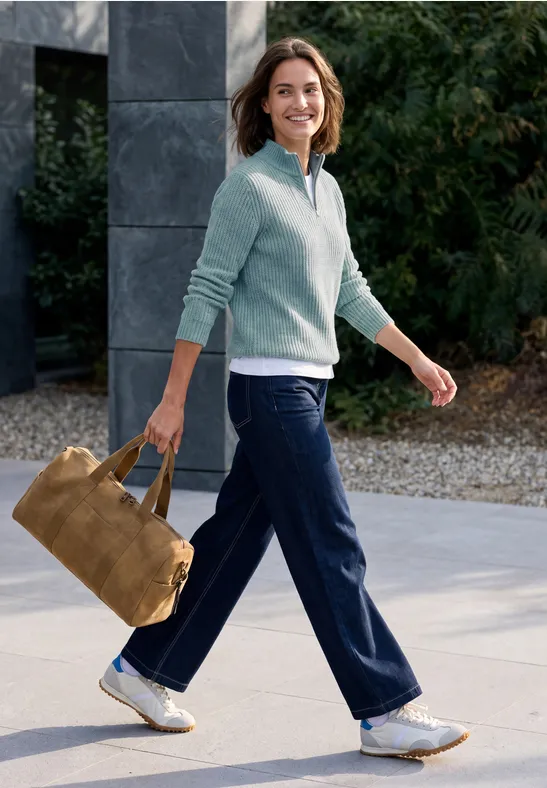 Frau spaziert draußen, lächelnd, trägt blaue Hose, hellblauen Pullover und eine braune Wildledertasche.