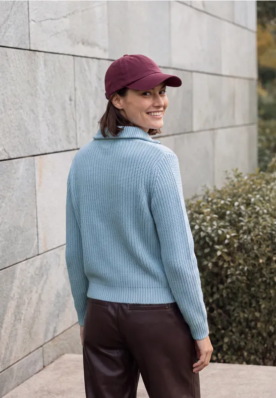 Frau in blauem Pullover und roter Cap steht seitlich an einer Mauer, blickt lächelnd über die Schulter.