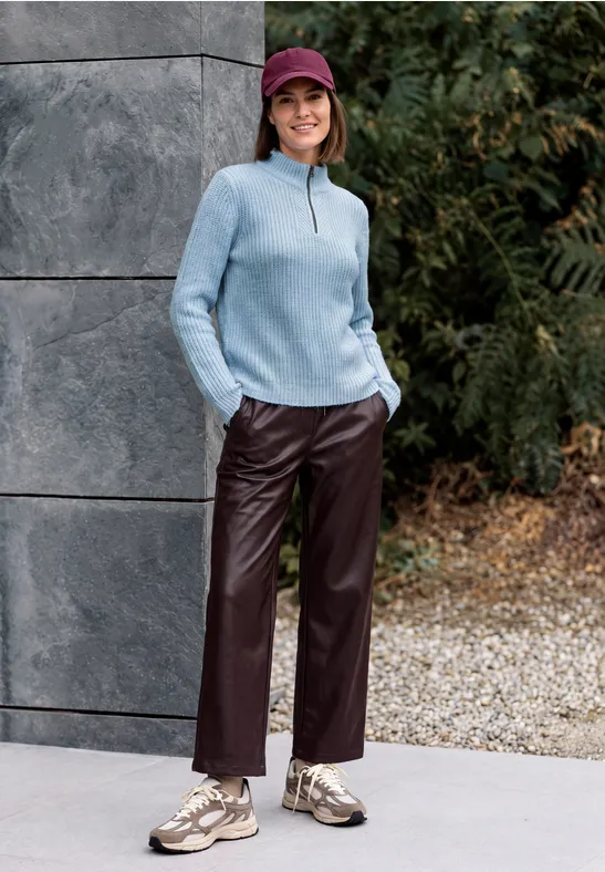 Eine Frau in blauem Pullover, roter Cap und Lederhose lehnt lächelnd an einer Mauer im Freien.