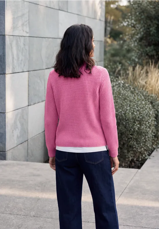 Frau in pinkem Pullover und dunkler Jeans steht im Freien, Rücken zur Kamera, vor Mauer und Büschen.