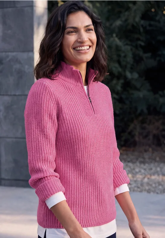 Frau steht lächelnd im Freien, trägt pinken Pullover vor ruhigem, grauem Hintergrund.