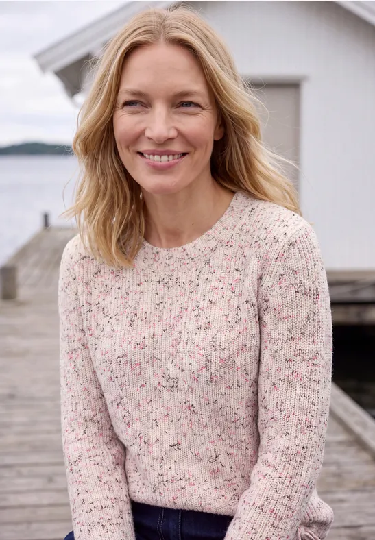 Frau mit blondem Haar lächelt auf einem Steg vor einem weißen Haus am Wasser.