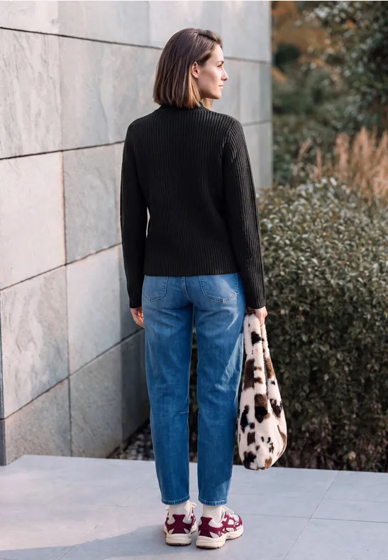 Frau von hinten, trägt schwarzen Pullover, blaue Jeans und bunte Sneakers, steht neben einer Mauer, hält eine Tasche.