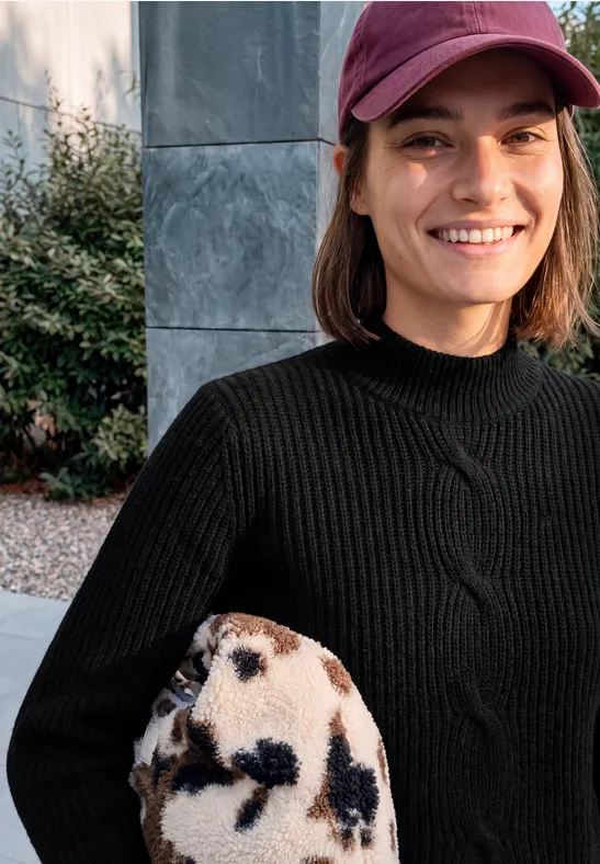 Lächelnde Frau in schwarzem Strickpullover und rosa Cap hält eine beige-schwarz gefleckte Felltasche.