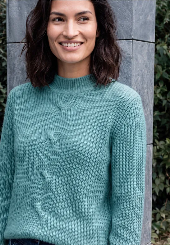 Eine lächelnde Frau in mintgrünem Strickpullover vor einer grauen Steinwand und Grünpflanzen.