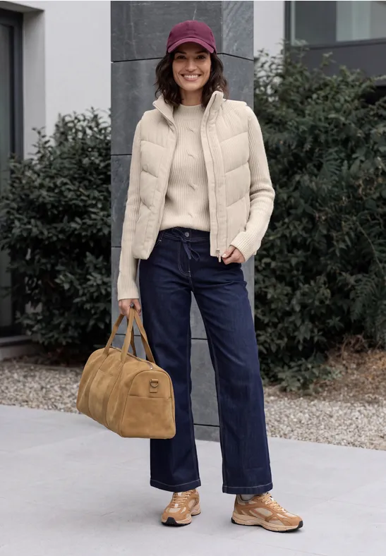 Vrouw met paarse pet, beige bodywarmer, trui, wijde jeans, bruine schoenen en tas voor een gebouw.