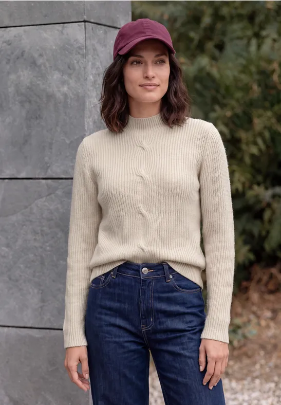 Vrouw met rode pet, beige trui en blauwe jeans voor een stenen muur in de buitenlucht.