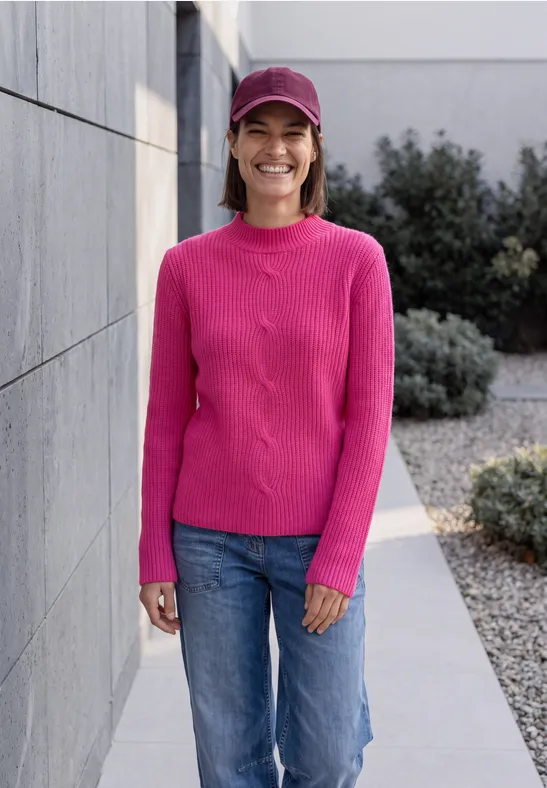 Fröhliche Frau in rosa Pullover, Jeans und Schirmmütze steht lächelnd vor einer Mauer und Buschwerk.