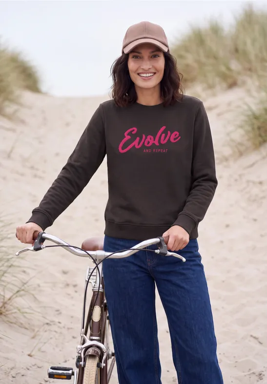 Frau mit Basecap und dunklem 'Evolve'-Sweatshirt lehnt am Fahrrad auf sandigem Dünenpfad