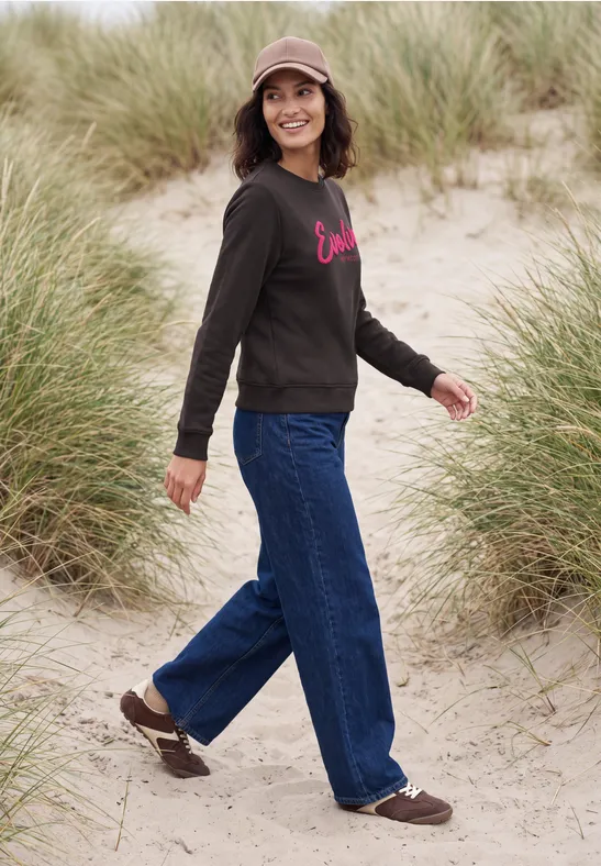 Eine Person spaziert über Sanddünen mit Dünengras, trägt Jeans, Sweatshirt, Mütze und braune Schuhe.