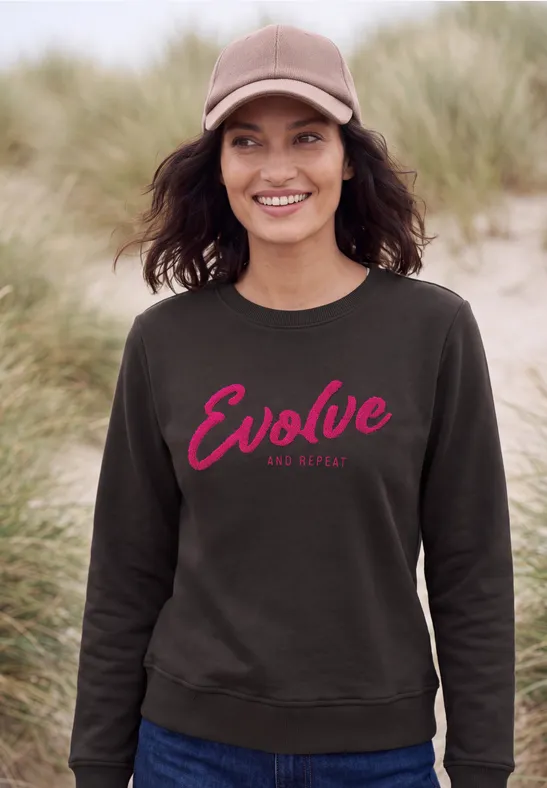 Frau in grauem Sweatshirt mit rotem 'Evolve'-Schriftzug an einem Dünenstrand.