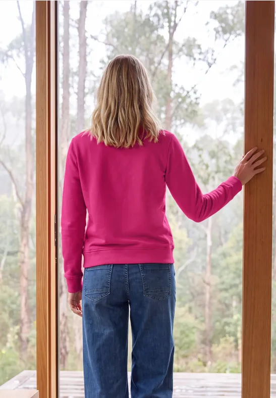 Person steht mit pinkem Pullover und Jeans im Türrahmen und blickt auf den Wald.