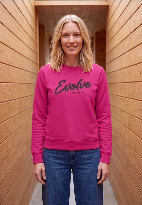 Eine lächelnde Frau in pinkem Sweatshirt mit 'Evolve'-Aufschrift steht in einem holzverkleideten Gang.
