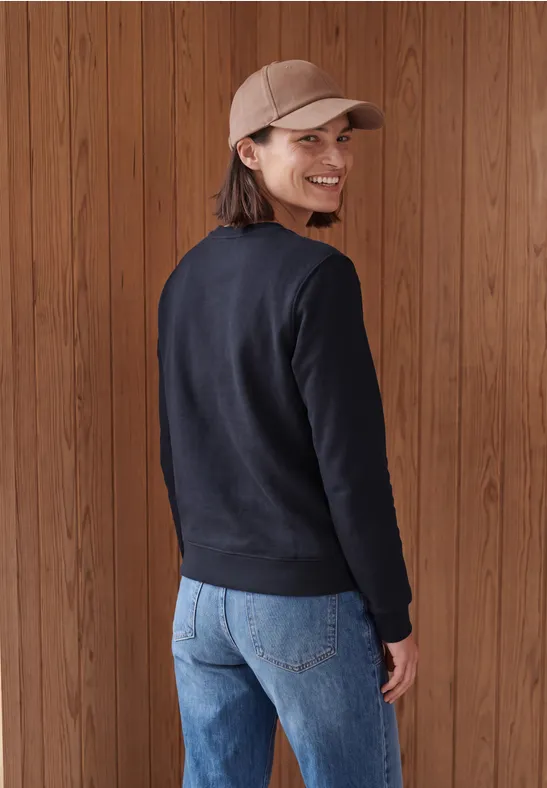Eine Frau mit Basecap, schwarzem Pullover und Jeans steht vor Holzpaneelen, blickt lächelnd über die Schulter.