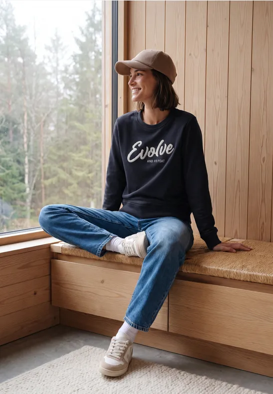 Frau sitzt gemütlich auf Fensterbank in Holzhütte, trägt Jeans, Sneaker und Cap, Pullover mit „Evolve“-Schriftzug.