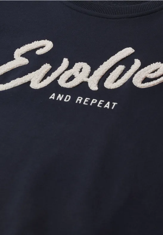Dunkelblaues T-Shirt mit weißem Schriftzug "Evolve and Repeat" in auffälliger, flauschiger Schrift.