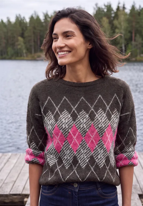 Eine lächelnde Frau in einem braunen Pullover mit pinkem Rautenmuster steht auf einem Steg vor einem See.
