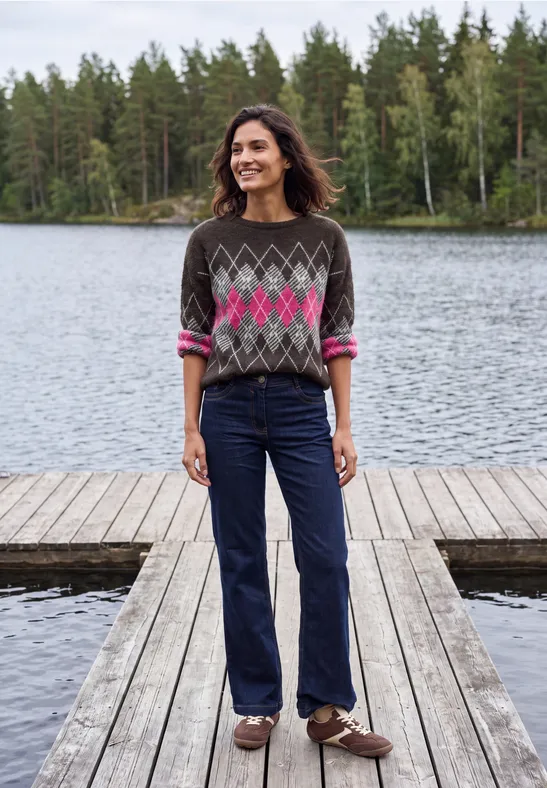 Eine lächelnde Frau steht auf einem Holzsteg am See vor Waldkulisse, sie trägt gemusterten Pullover und Jeans.