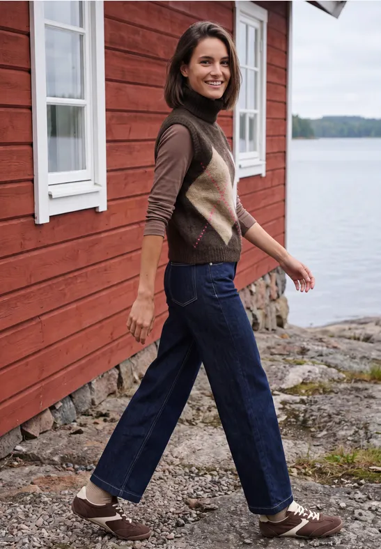Eine lächelnde Frau in Jeans und Weste spaziert an einem roten Holzhaus am Wasser.