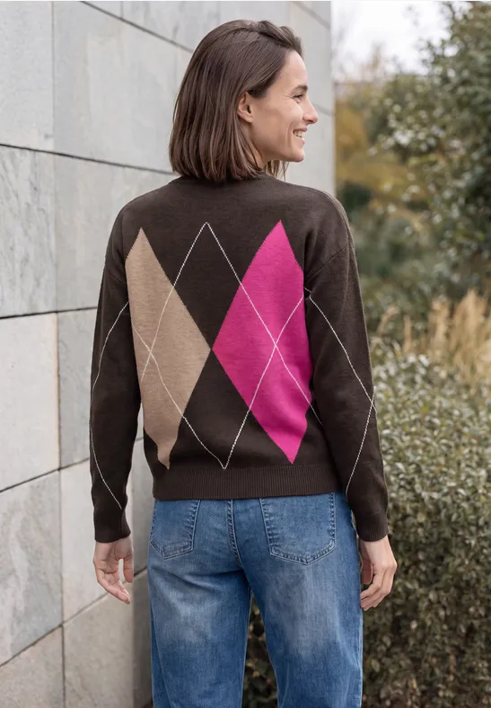 Frau in Jeans und braun-pinkem Rautenpullover, lächelnd in die Umgebung blickend.