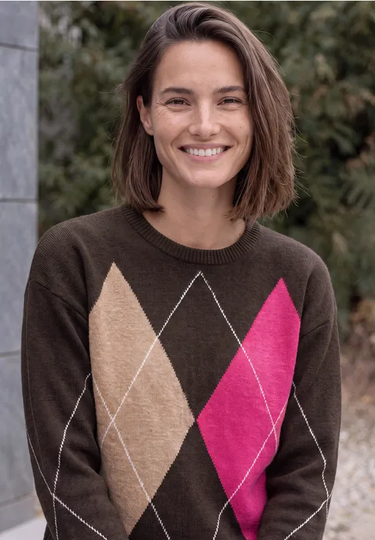 Lächelnde Frau mit kurzen braunen Haaren im braunen Argyle-Pullover mit beigen und pinken Rauten vor Büschen.
