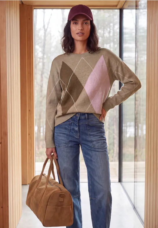 Frau mit roter Kappe, beigem Rautenpullover, blauen Jeans und brauner Tasche in hellem Raum vor Fenster.