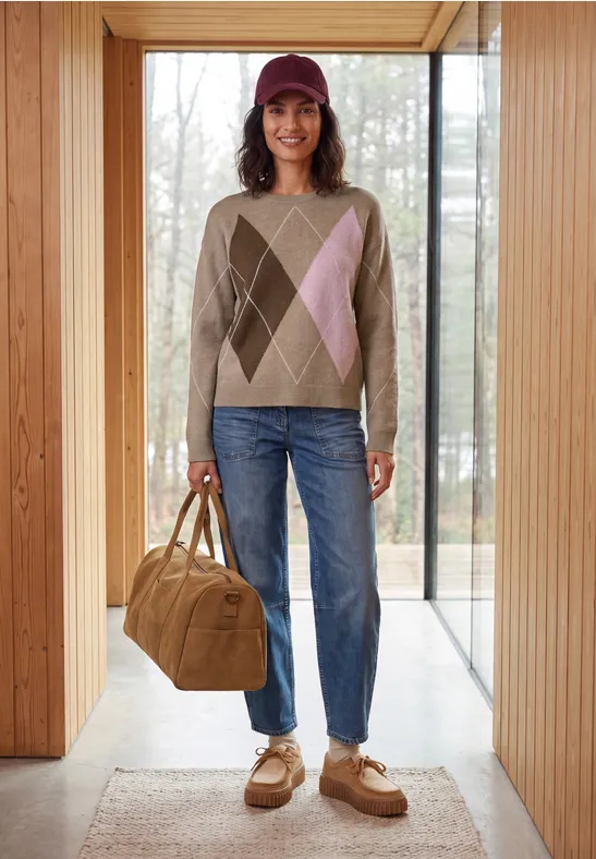 Frau steht in einer Holzrahmen-Tür, trägt argyle Pullover, Jeans, Cap und hält eine große Umhängetasche.