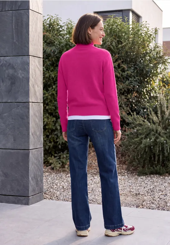 Een vrouw in roze trui en spijkerbroek staat buiten voor een huis en kijkt naar rechts.