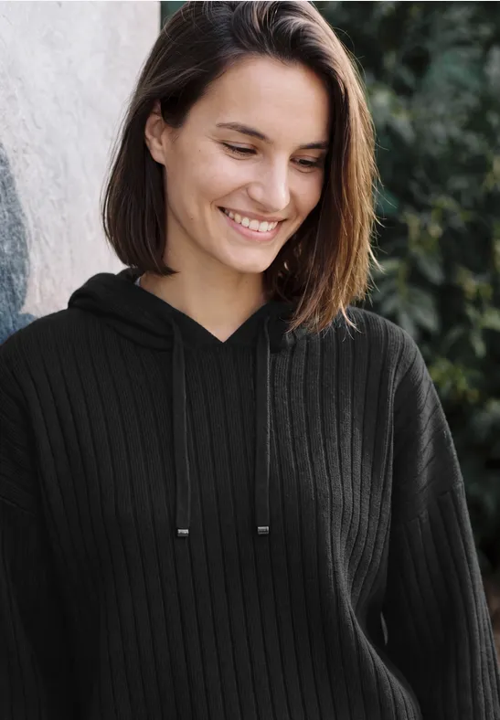 Eine lächelnde junge Frau in schwarzem Pullover mit Kapuze vor grünem Hintergrund.
