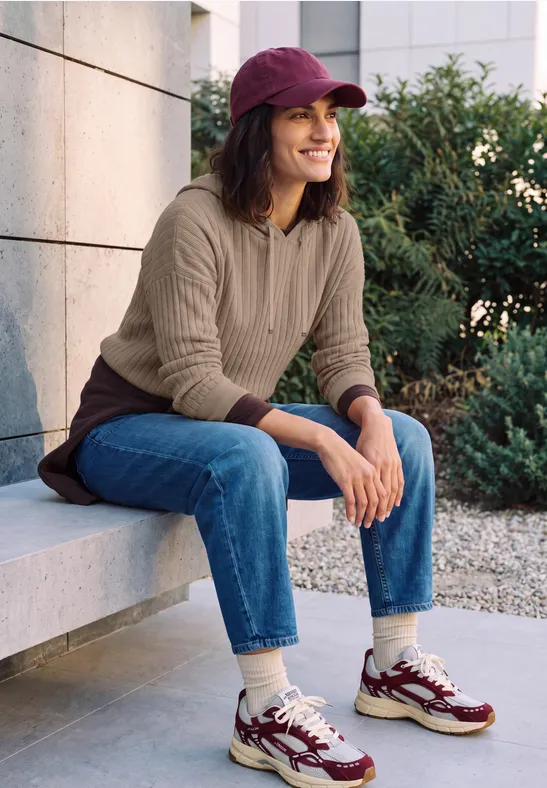 Femme à casquette rouge, pull beige, jean et baskets, assise et souriante sur un muret en plein air.