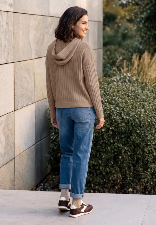 Femme en sweat à capuche marron, jean retroussé et baskets, appuyée contre un mur avec des plantes derrière elle.