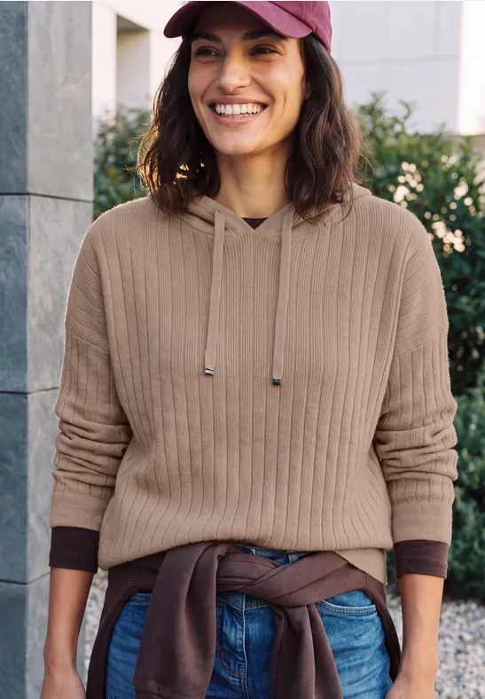 Femme souriante en pull beige, jean et casquette devant un mur urbain.